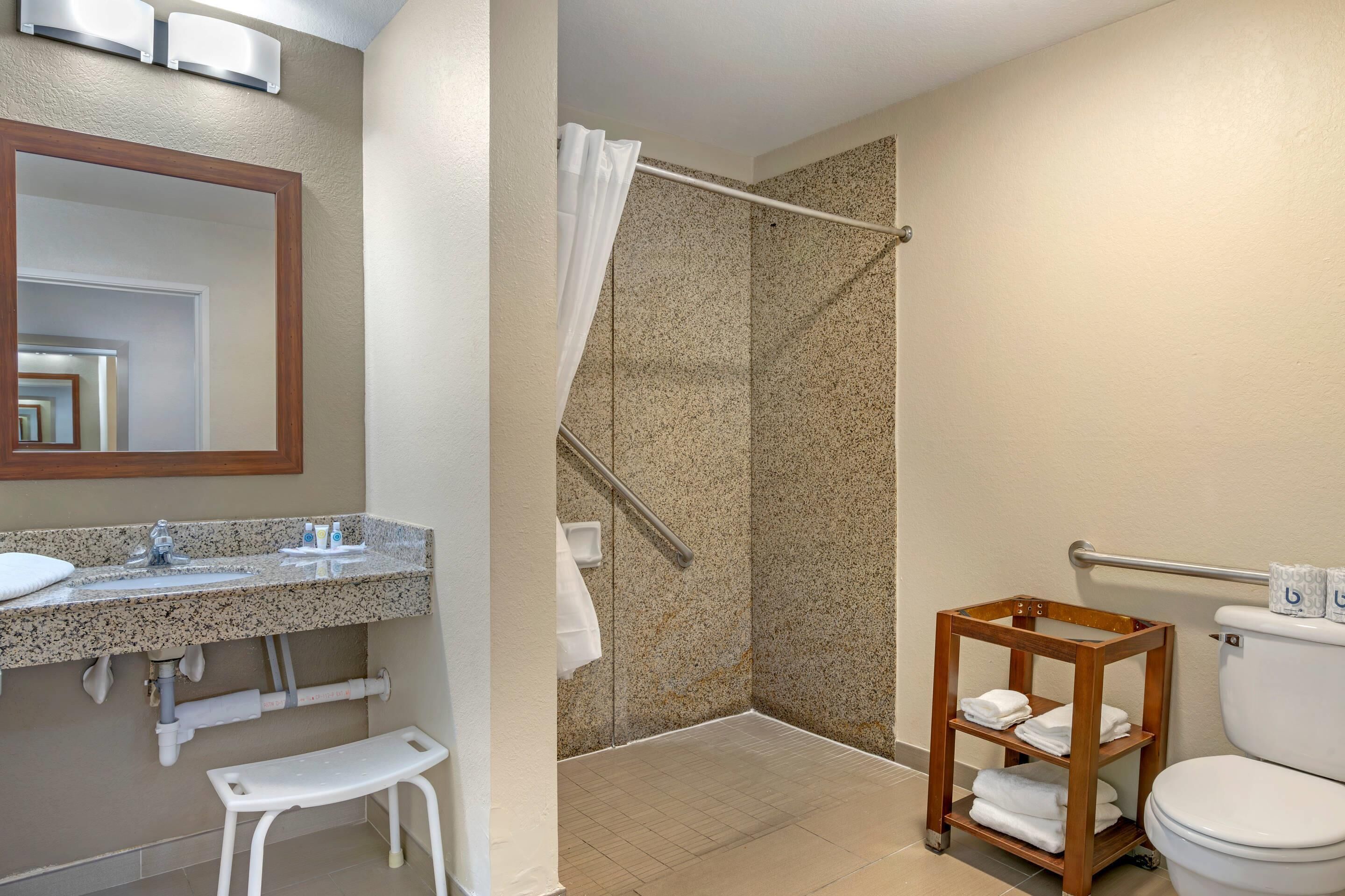 Une salle de bain avec un lavabo sur un plan de travail en granit, un miroir, une douche avec un rideau, une étagère en bois avec des serviettes et des toilettes.