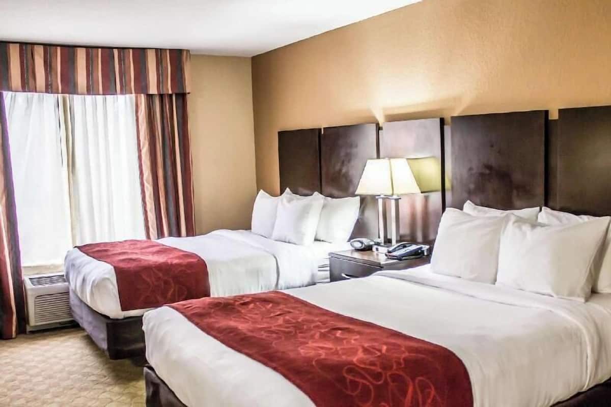 A hotel room with two beds, a headboard, a nightstand with a lamp, and a window with curtains.