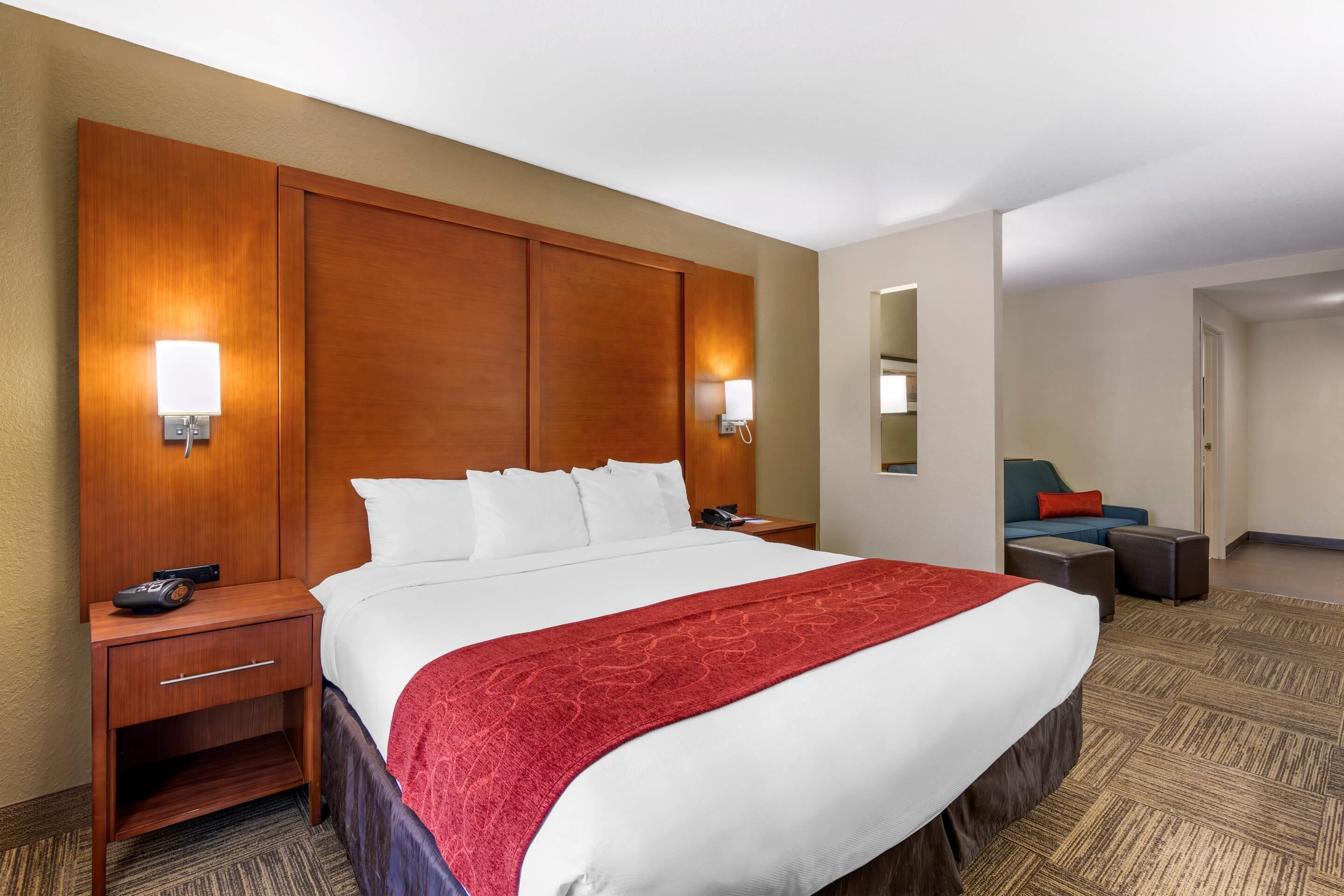 Ein Hotelzimmer mit einem großen Bett, einem Holzkopfende, Nachttischen mit Lampen, einer roten Tagesdecke und einer Sitzecke mit Sofa und Hocker.