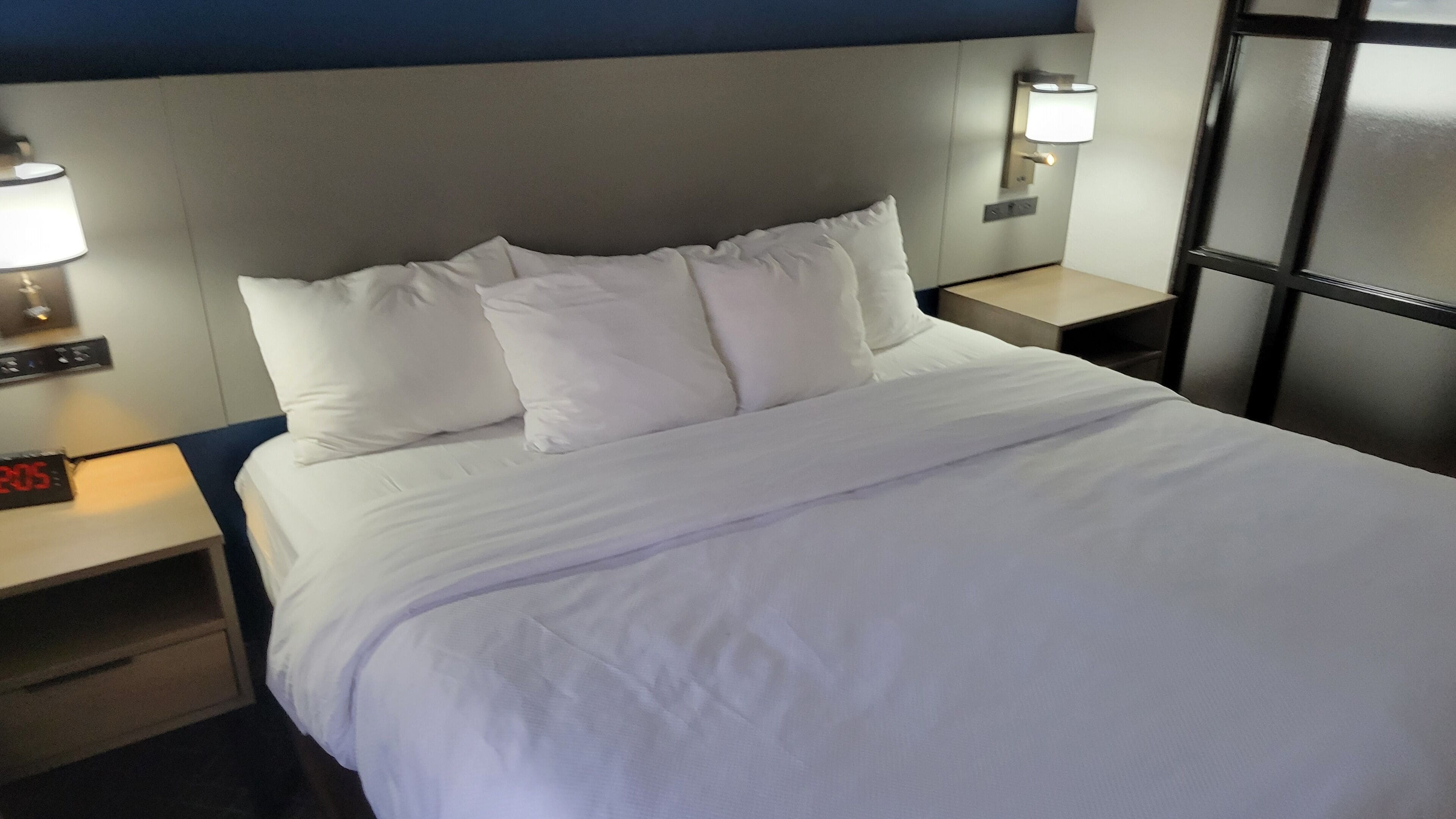 A neatly made bed with white linens, a digital clock, and a wall-mounted lamp.