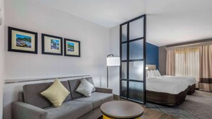 A hotel room with a grey sofa, a yellow side table, a bed with two pillows, and a large window with curtains. - Comfort Suites Macon Northwest (Macon)