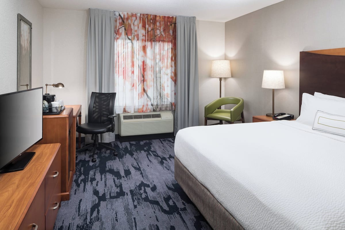 Habitación de hotel con cama, escritorio con televisor, silla, lámpara y ventana con cortinas.