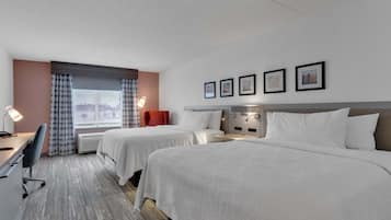 Quarto de hotel com duas camas, uma escrivaninha, uma cadeira, janela com cortinas e quatro quadros emoldurados na parede.