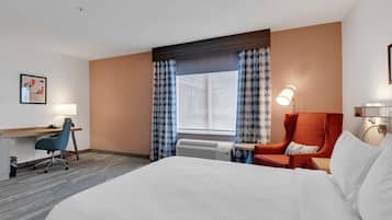 A hotel room with a large bed, a desk with a chair, a red armchair, a window with curtains, and a wall-mounted lamp.