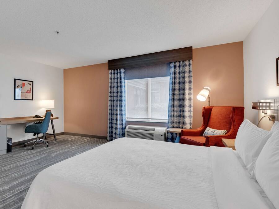 A hotel room with a large bed, a desk with a chair, a red armchair, a window with curtains, and a wall-mounted lamp.
