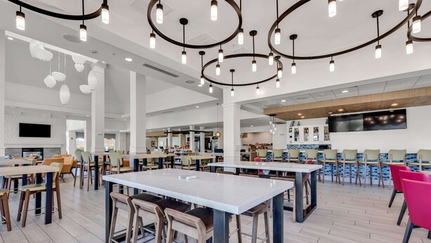 A spacious dining area with multiple tables and chairs, a modern ceiling design with hanging lights, and a large flat-screen TV mounted on the wall. - Hilton Garden Inn Myrtle Beach/Coastal Grand Mall (Myrtle Beach)
