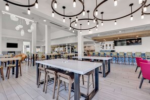 A spacious dining area with multiple tables and chairs, a modern ceiling design with hanging lights, and a large flat-screen TV mounted on the wall. - Hilton Garden Inn Myrtle Beach/Coastal Grand Mall (Myrtle Beach)