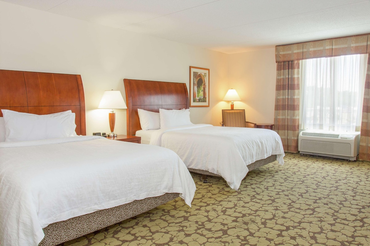 A hotel room with two beds, a wooden headboard, a framed picture on the wall, a lamp, and a window with curtains.