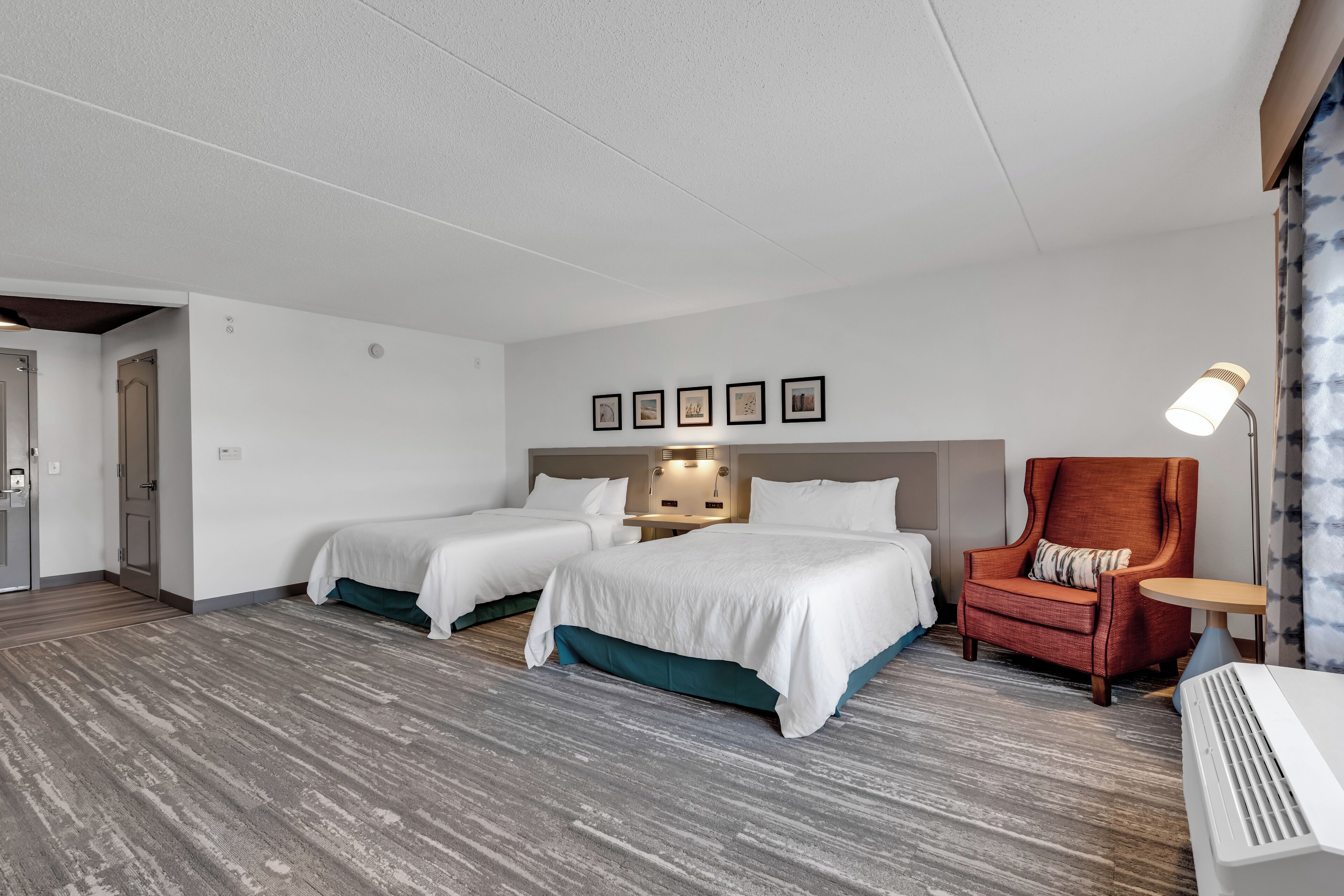 A hotel room with two beds, a red armchair, a small table, and a wall-mounted lamp.