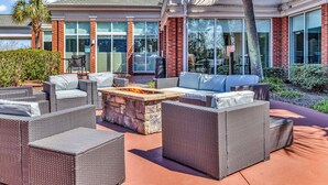 An outdoor patio with wicker furniture, a stone fire pit, and a view of a building with large windows and a brick facade. - Hilton Garden Inn Myrtle Beach/Coastal Grand Mall (Myrtle Beach)
