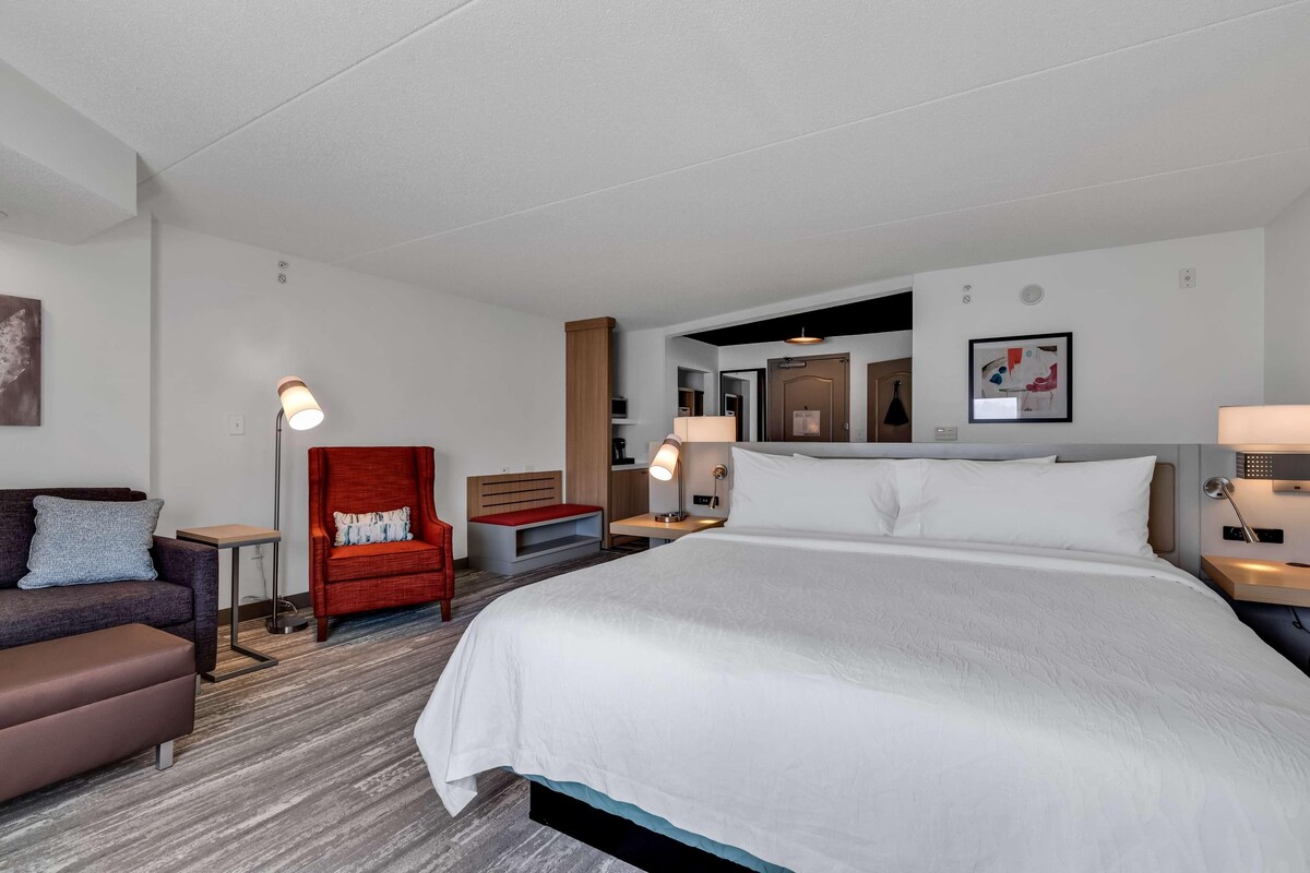 A hotel room with a large bed, a red armchair, a sofa, a desk, and a lamp.