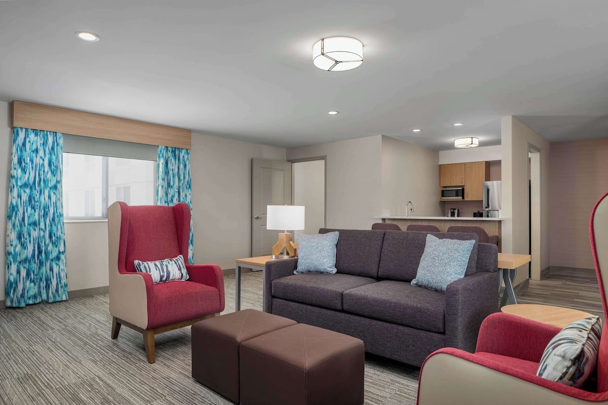 A hotel room with a sofa, armchair, ottoman, and a kitchenette in the background.