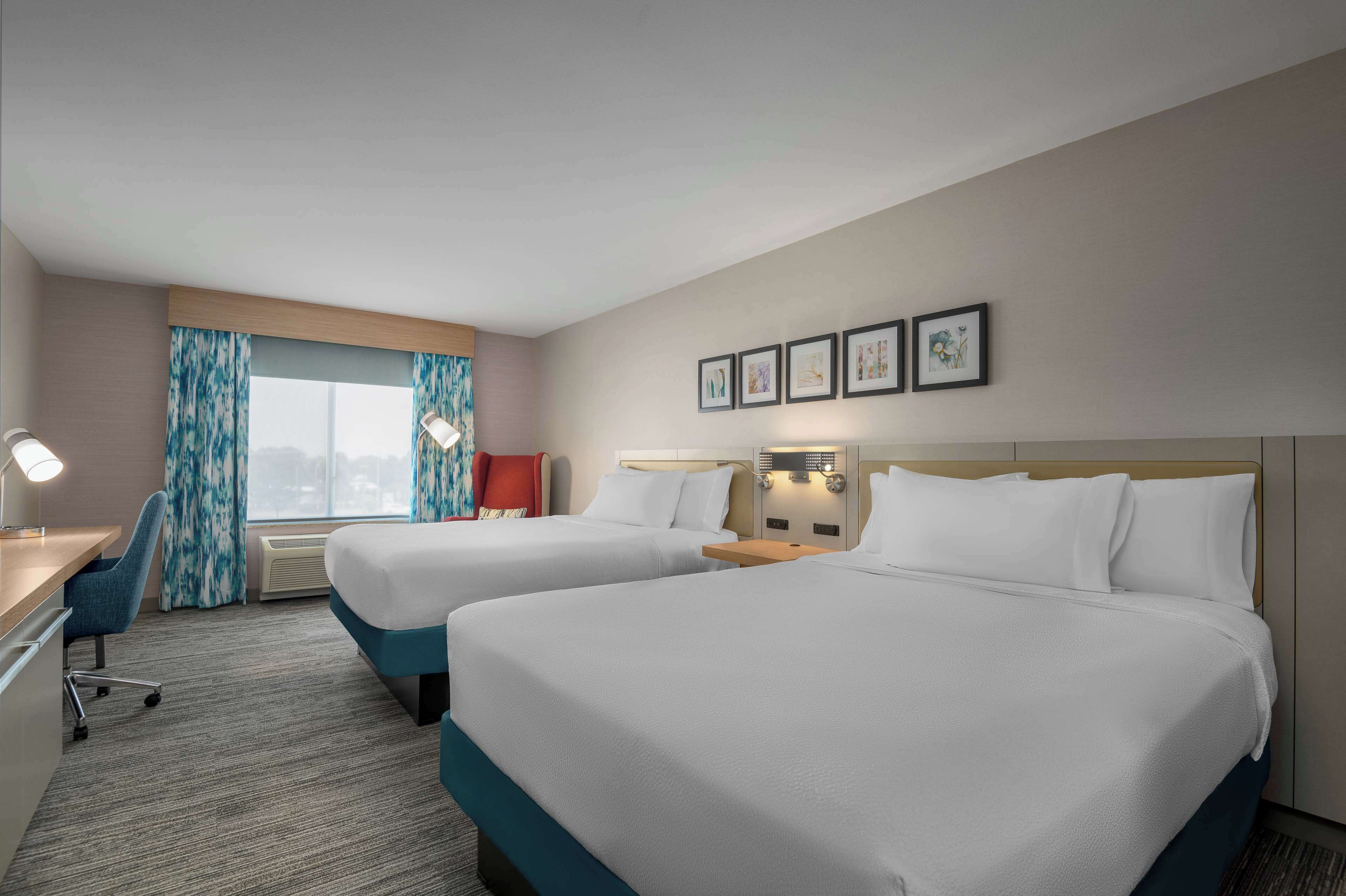 A hotel room with two beds, a desk, a chair, and a large window with curtains.