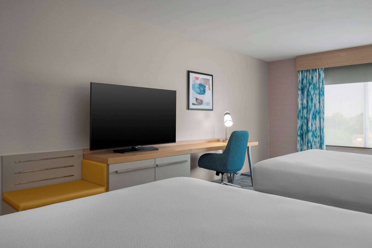 A hotel room with two beds, a desk with a flat-screen TV, a chair, and a window with curtains.