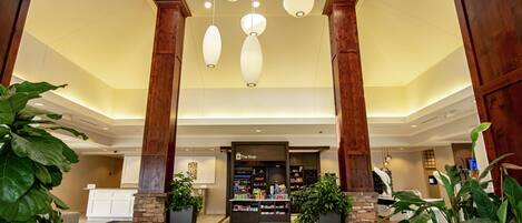 A hotel lobby with a reception desk, large potted plants, and a ceiling with hanging light fixtures.