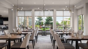 A modern dining area with large windows, multiple tables, and chairs, featuring a central chandelier and a view of the outdoors.