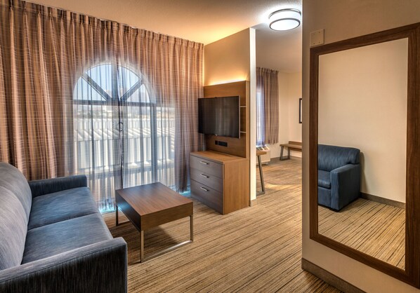 Una habitación de hotel moderna con un ventanal grande, un sofá, una mesa de centro, un televisor y un espejo.