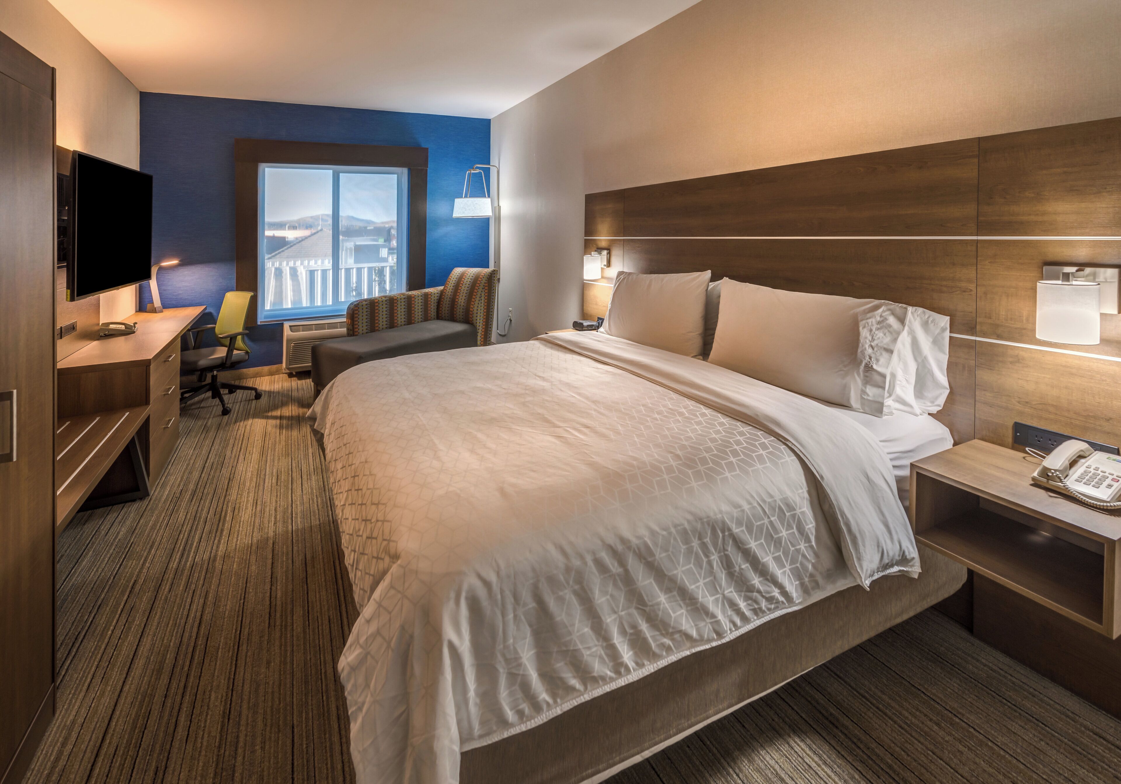 A hotel room with a large bed, a desk with a TV, a chair, a bedside table with a lamp, and a window with a view.