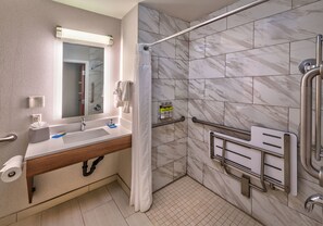Un baño moderno con ducha, barras de apoyo, lavamanos con espejo y dispensador de papel higiénico.