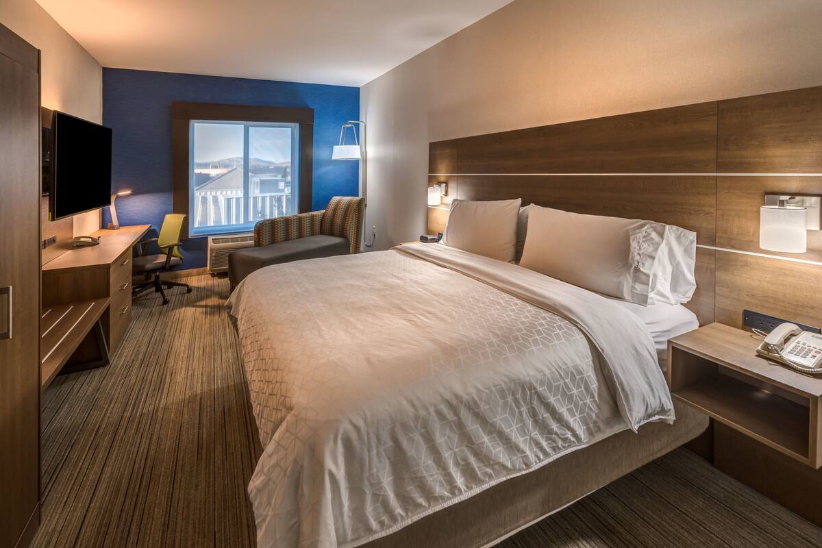 A hotel room with a large bed, a desk with a flat-screen TV, a chair, a bedside table with a lamp, and a window with a view of a building.