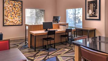 Habitación de hotel con escritorio, dos sillas, computadora e impresora. Se ven ventanas con vistas a árboles y un cuadro en la pared.