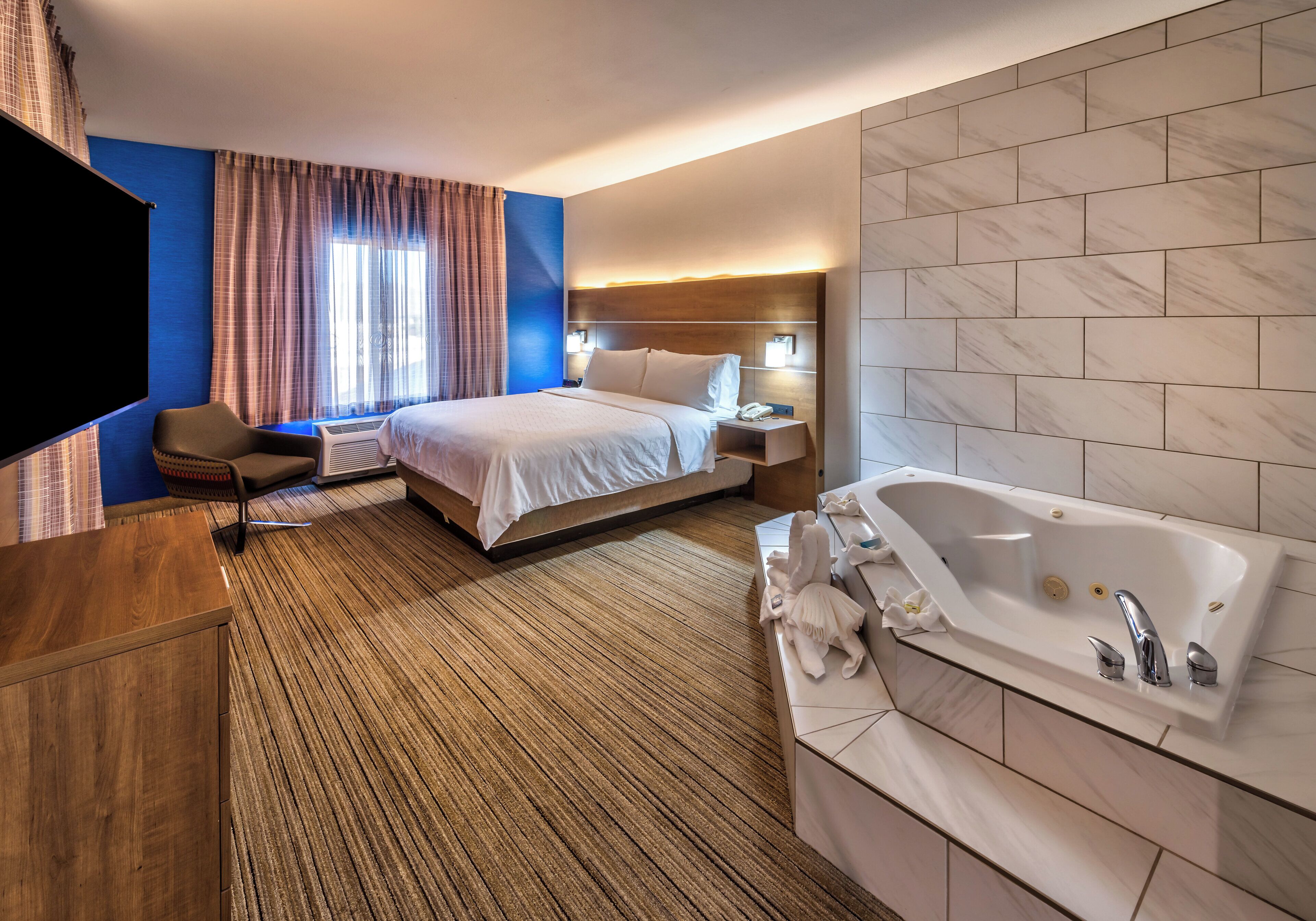 A hotel room with a bed, a large window with curtains, a flat-screen TV, a bedside table, a nightstand, a bathroom with a bathtub, and a marble wall.