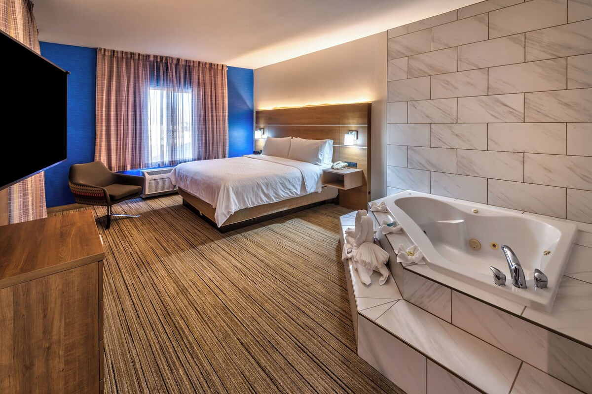 Habitación de hotel con cama, un ventanal con cortinas, televisor de pantalla plana, mesita de noche y baño con bañera, además de una pared de mármol.