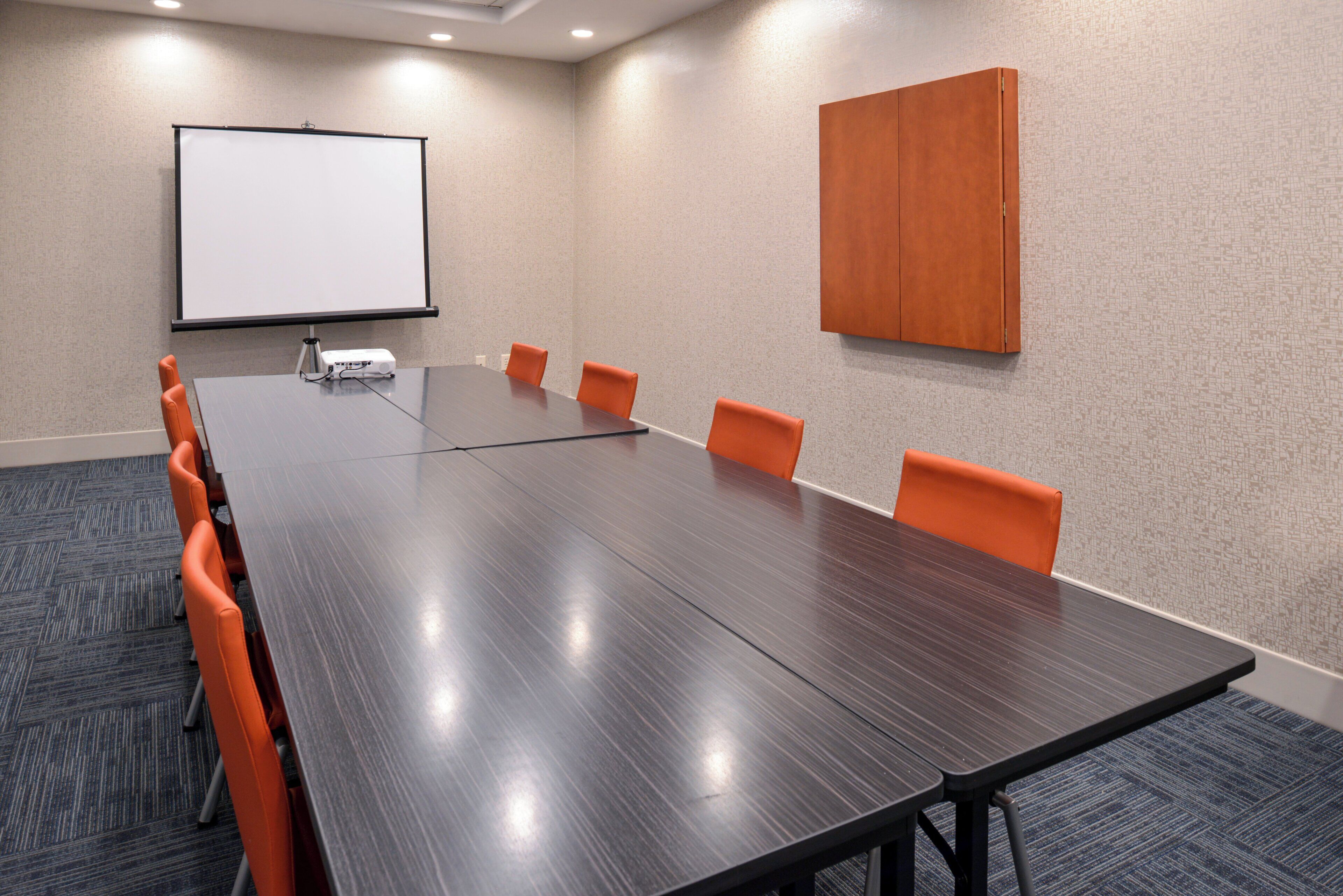 A conference room with a long table, six orange chairs, a projector, and a white screen on the wall.