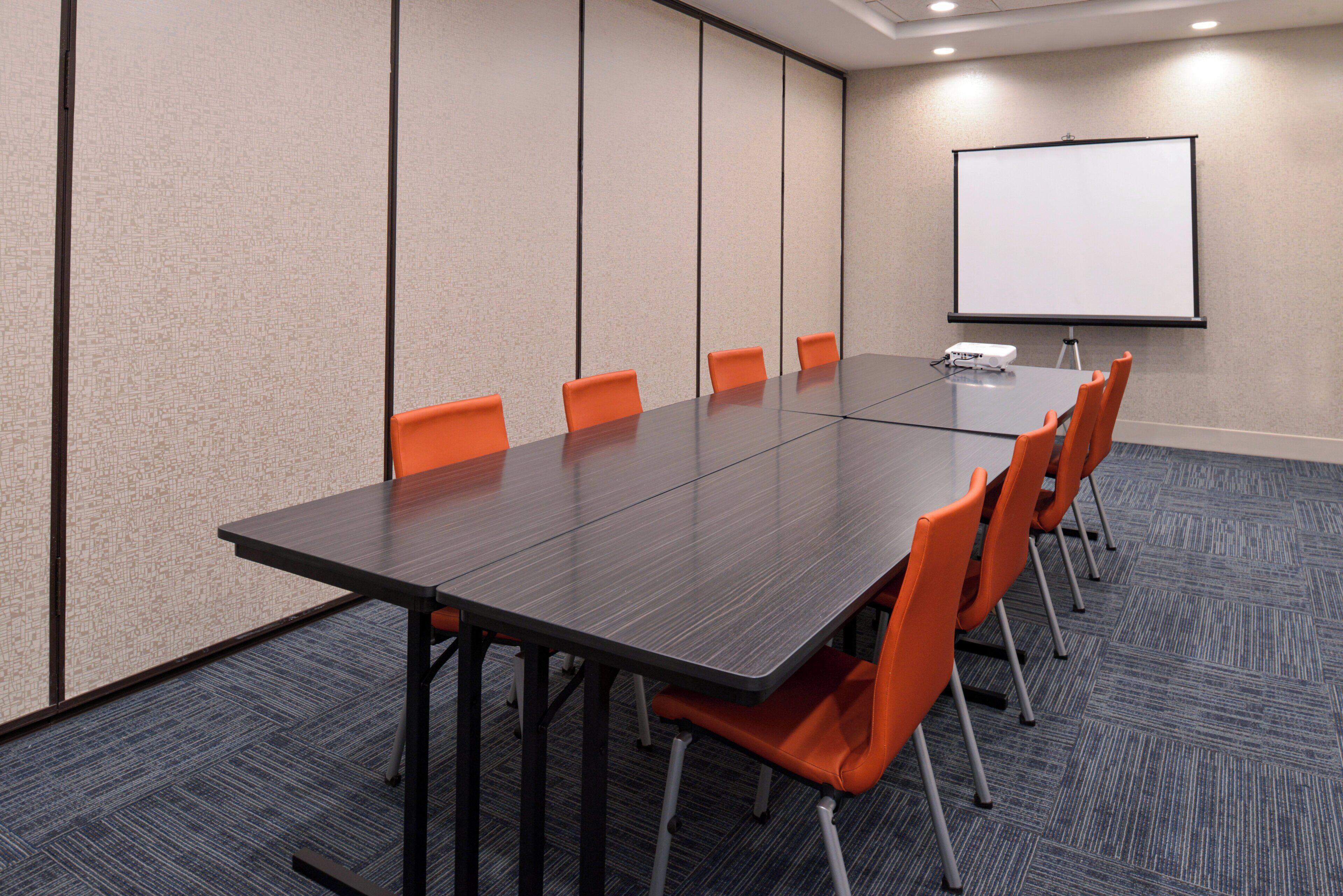 A conference room with a long table, six orange chairs, a projector, and a white screen.