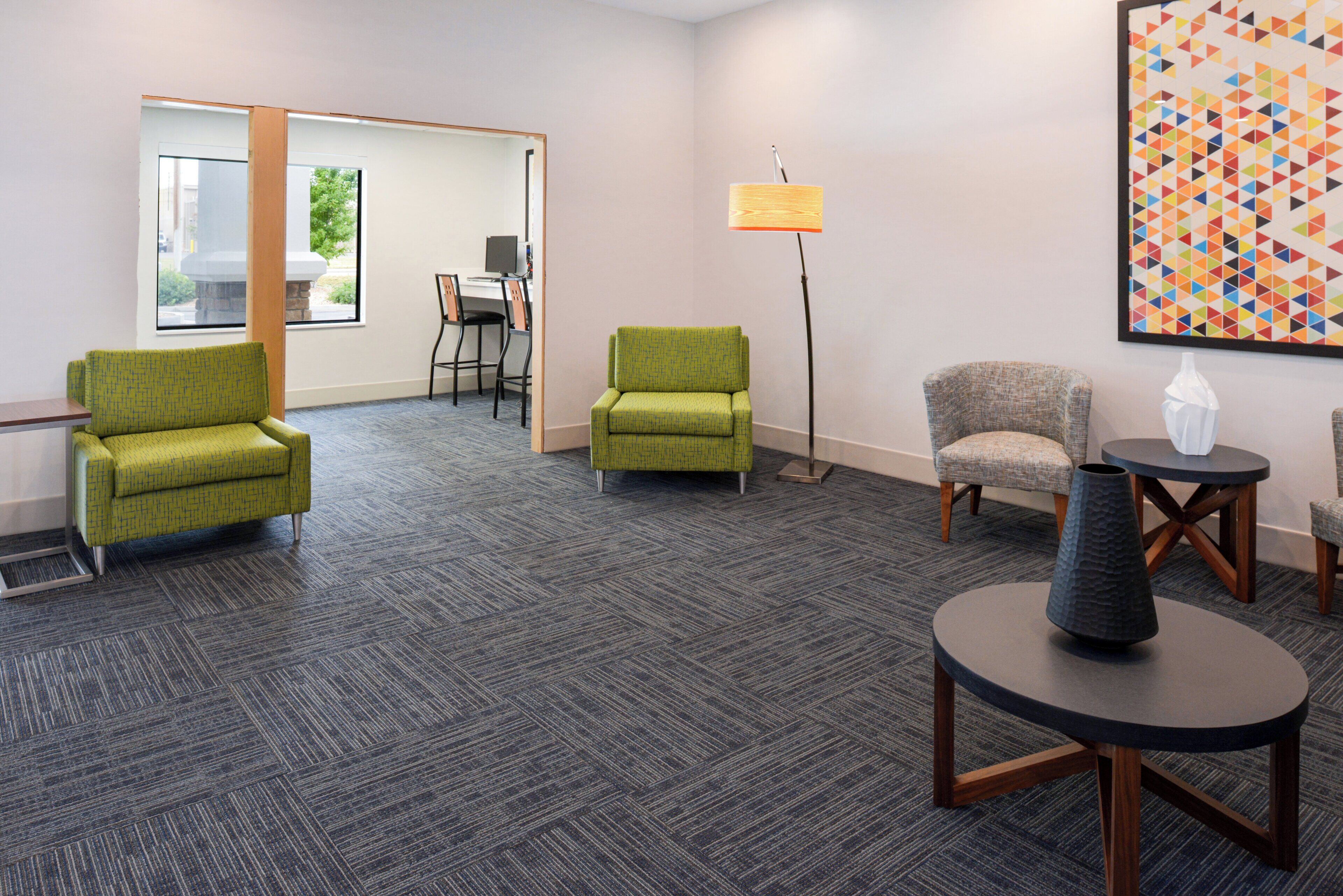 A hotel room with two green armchairs, a grey armchair, a floor lamp, a round table with a vase, and a large geometric painting on the wall.