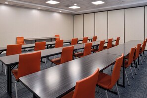 Een vergaderzaal met meerdere tafels en stoelen, een vloerbedekking en een effen wand met een deur.