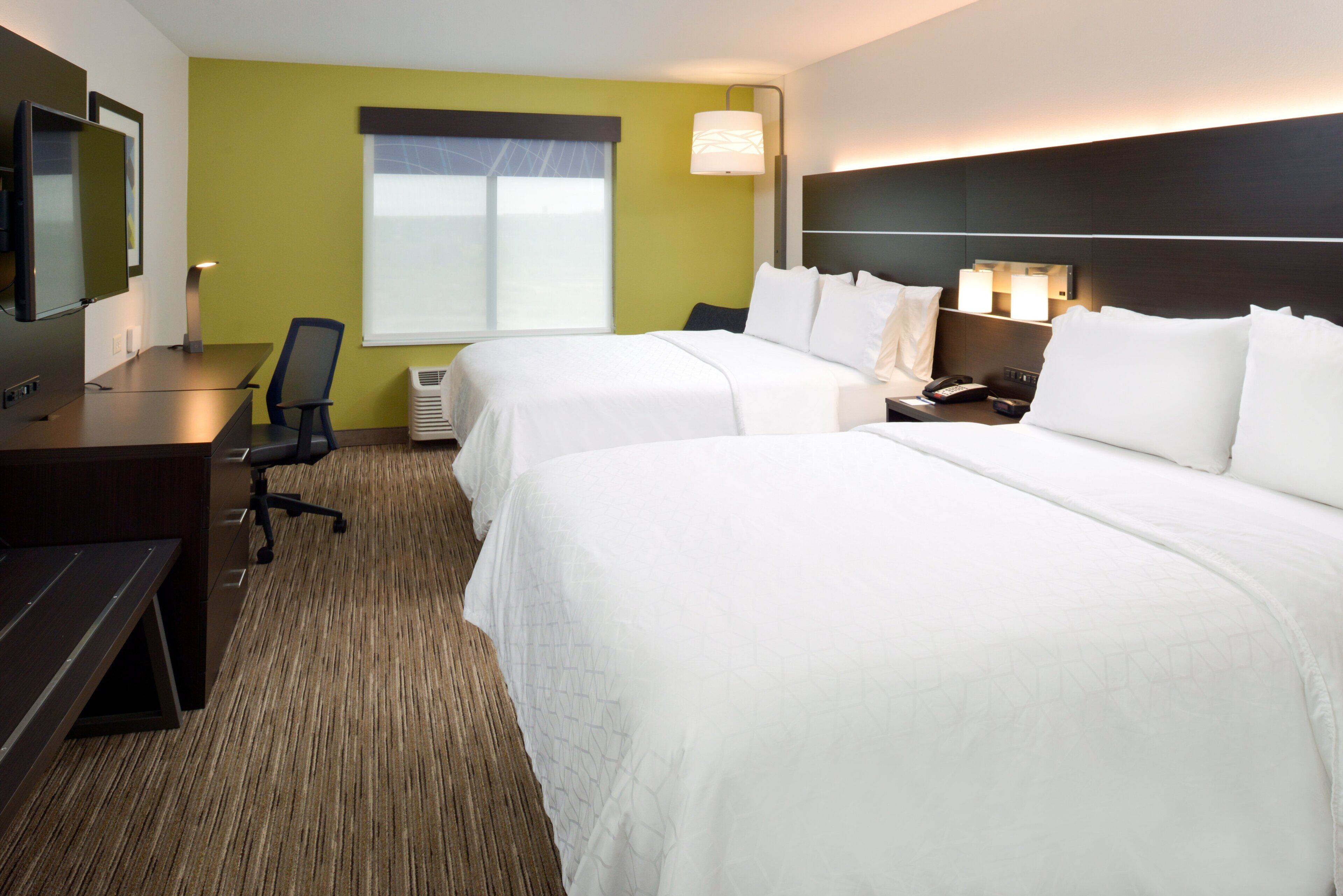 Une chambre d’hôtel avec deux lits, un bureau, une chaise, une télévision et une fenêtre avec des stores.
