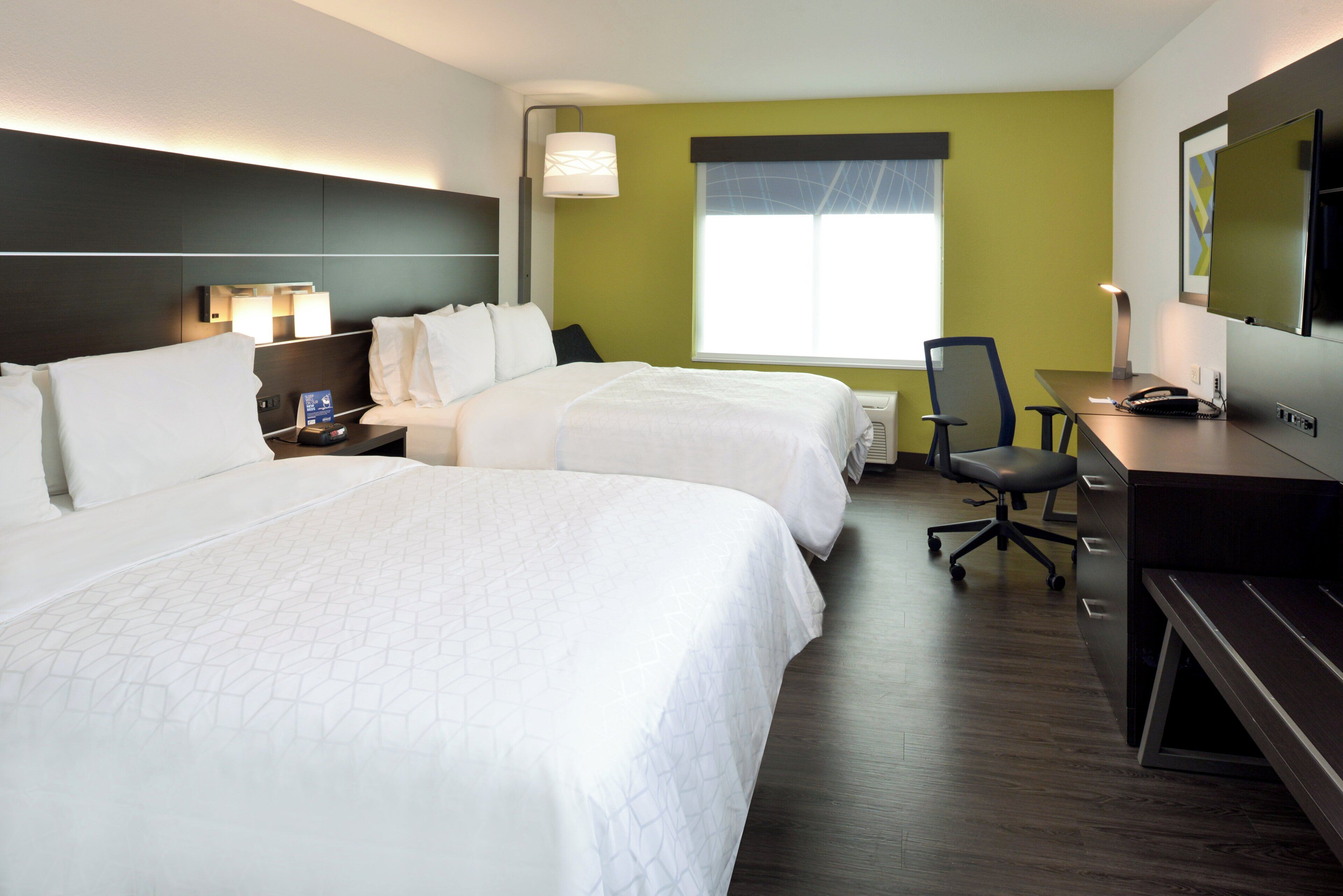 Quarto de hotel com duas camas, uma escrivaninha, uma cadeira, televisão e janela com persianas.