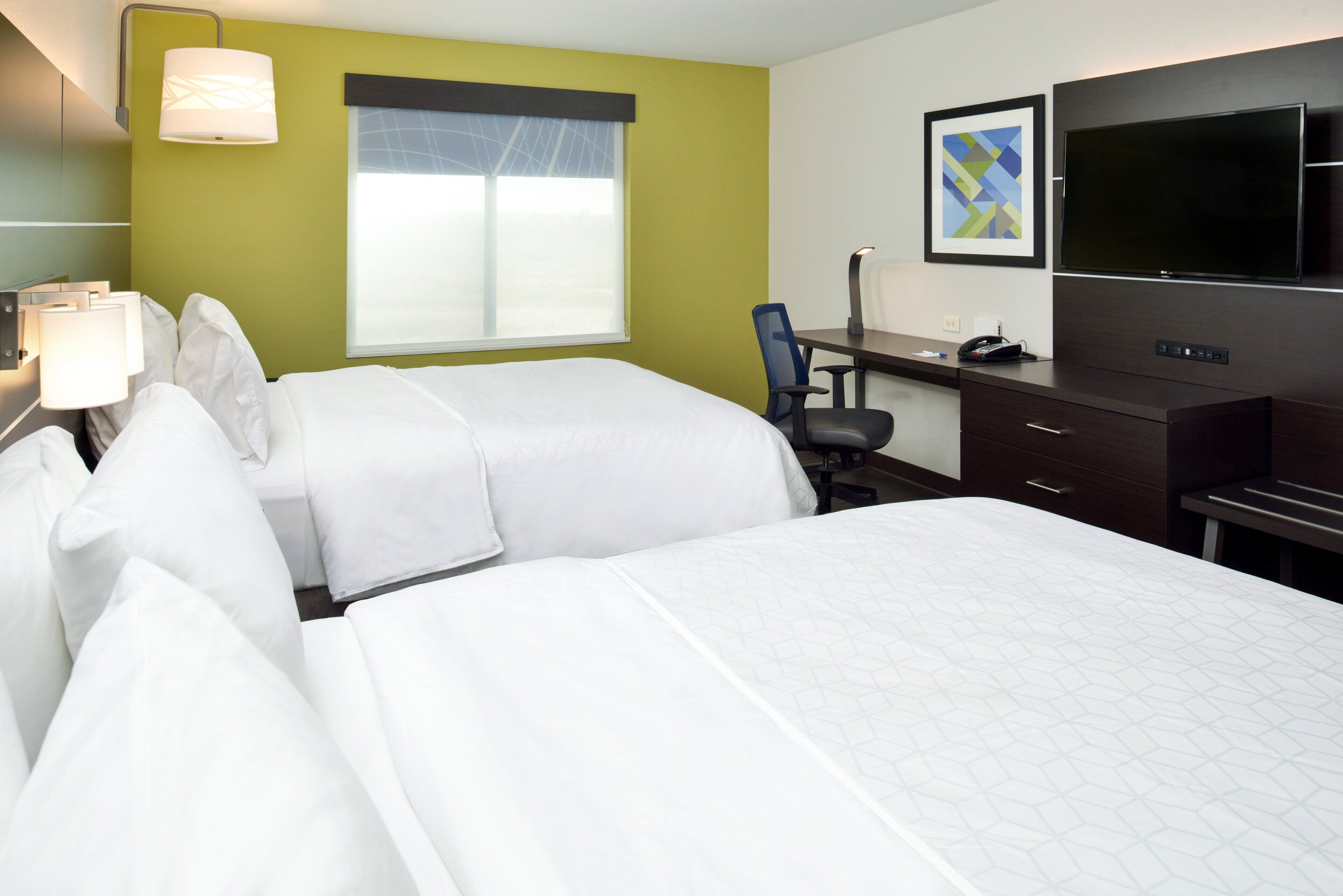 A hotel room with two beds, a desk with a chair, a TV, and a window with blinds.