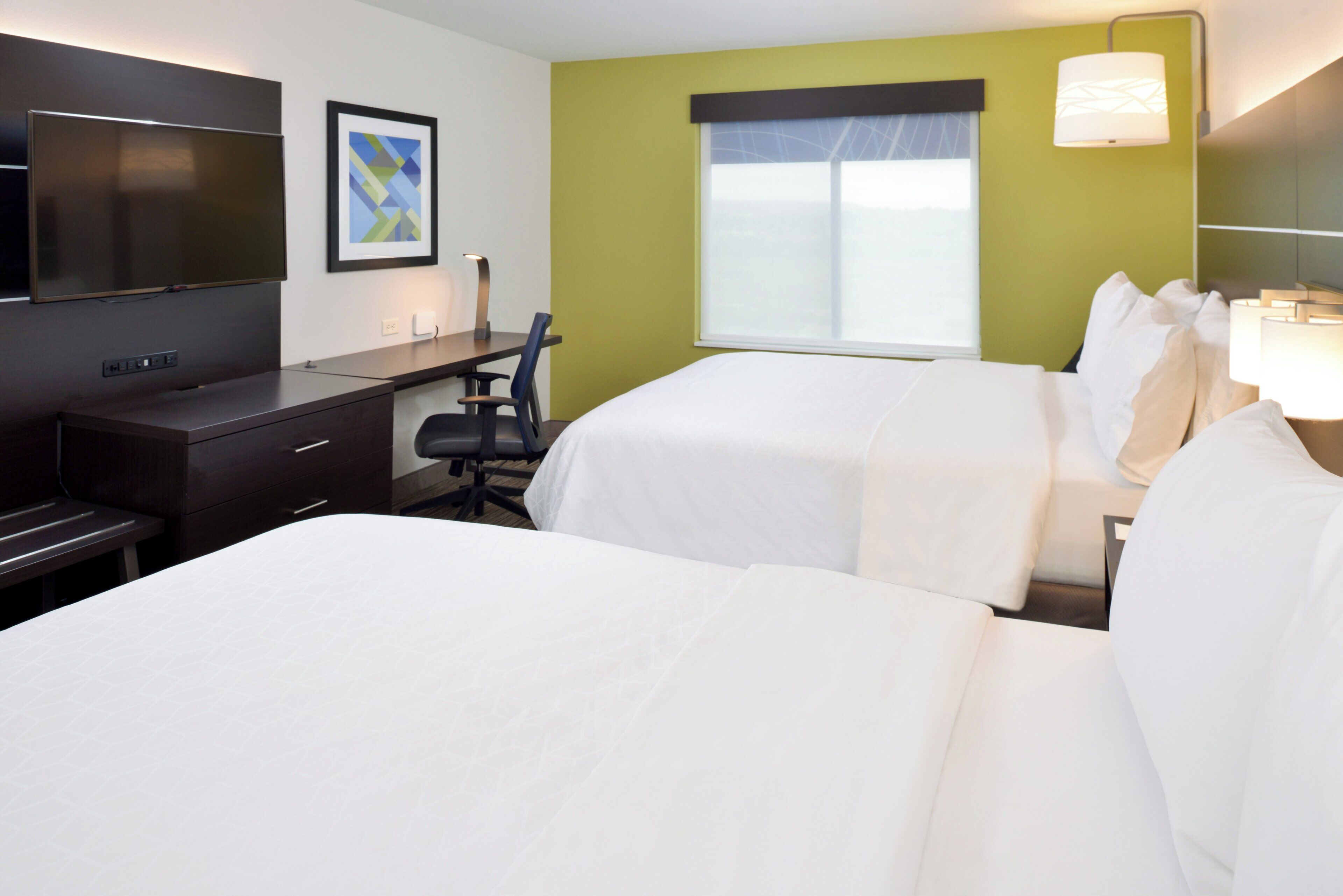 A hotel room with two beds, a desk, a chair, a TV, and a window with blinds.
