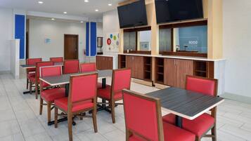 A modern dining area with red chairs and wooden tables, equipped with a flat-screen TV and a blue wall with a clock.