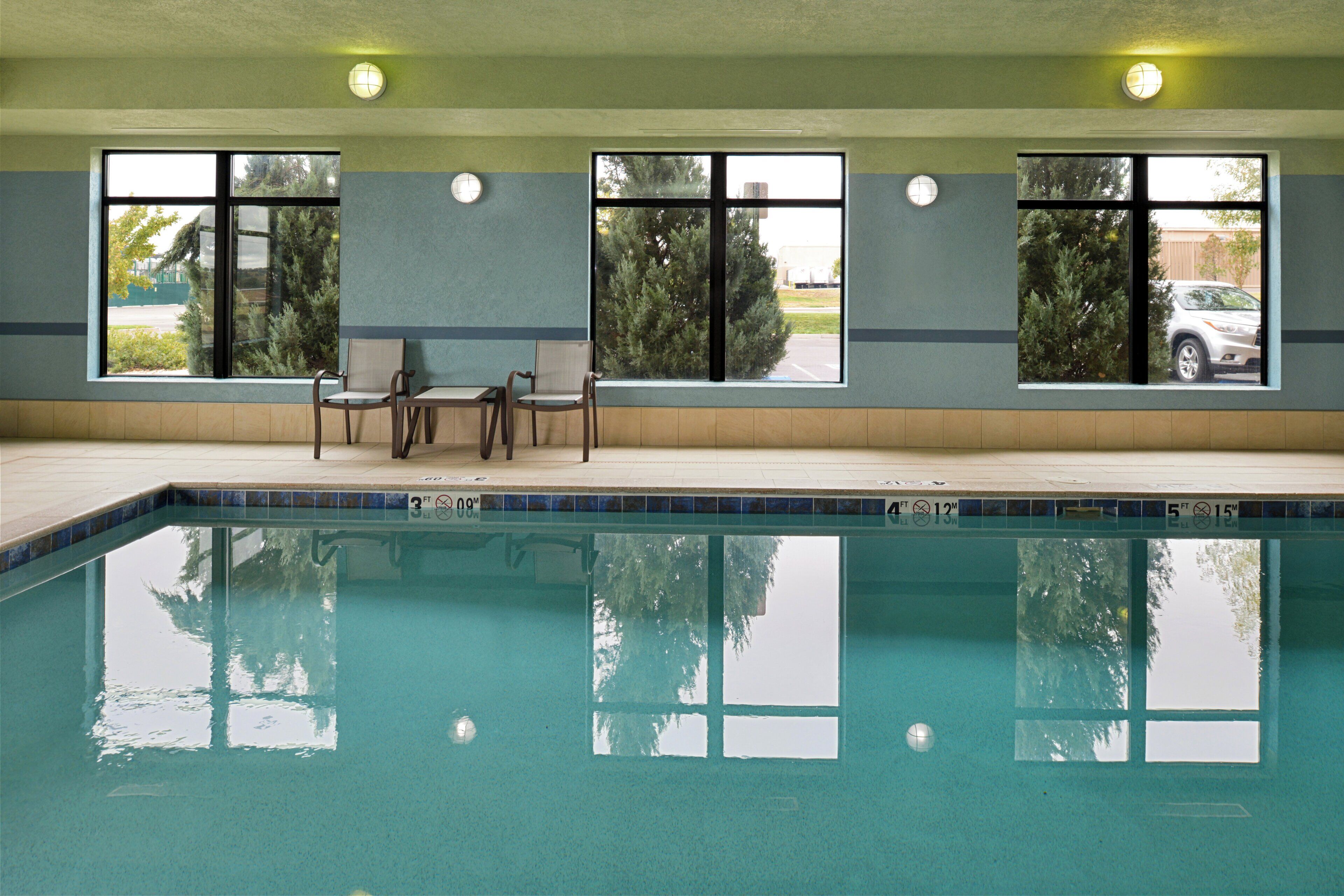 Une piscine avec un bord carrelé, un espace détente avec des chaises, et des fenêtres donnant sur des arbres et un bâtiment.