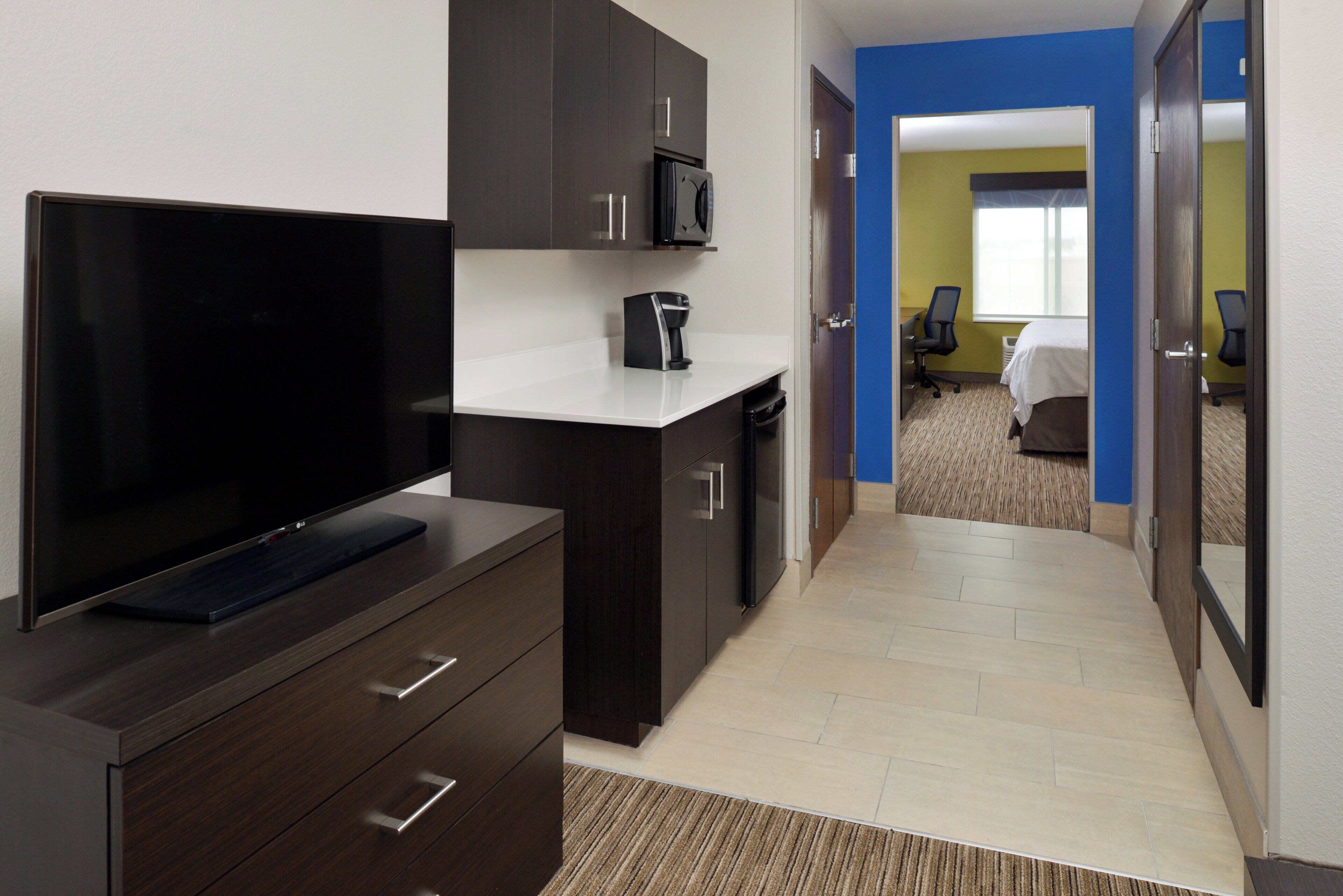 A hotel room with a flat-screen TV, dark wooden furniture, a white countertop, and a view into a room with a bed and a chair.