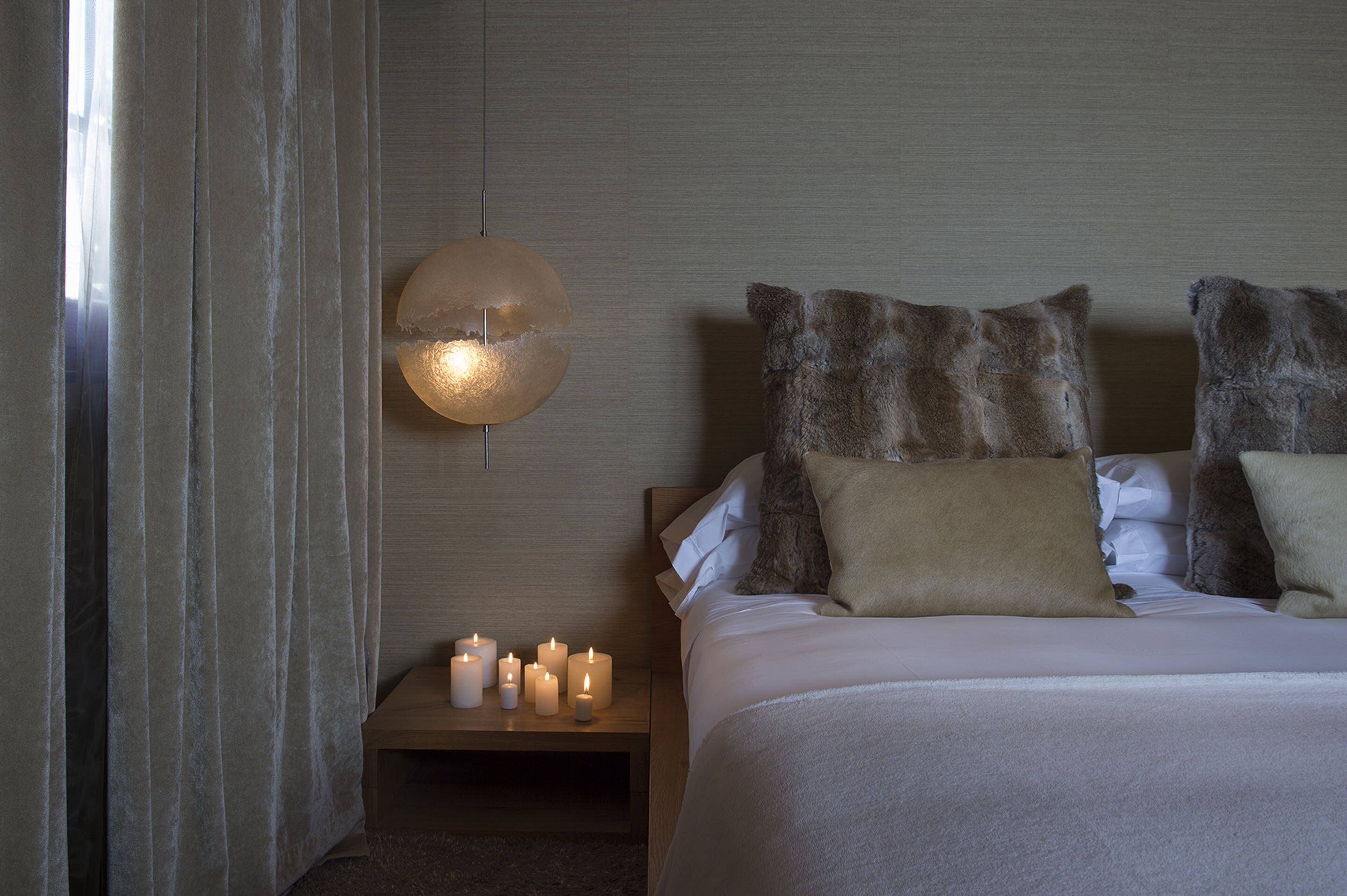 En pænt redt seng med mange puder, et natbord med tændte stearinlys og en kugleformet lampe på væggen.