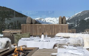 An outdoor dining area with a wooden table set for a meal, a fire pit, and a view of snow-capped mountains. - Grau Roig Andorra Boutique Hotel & Spa (Grau Roig)