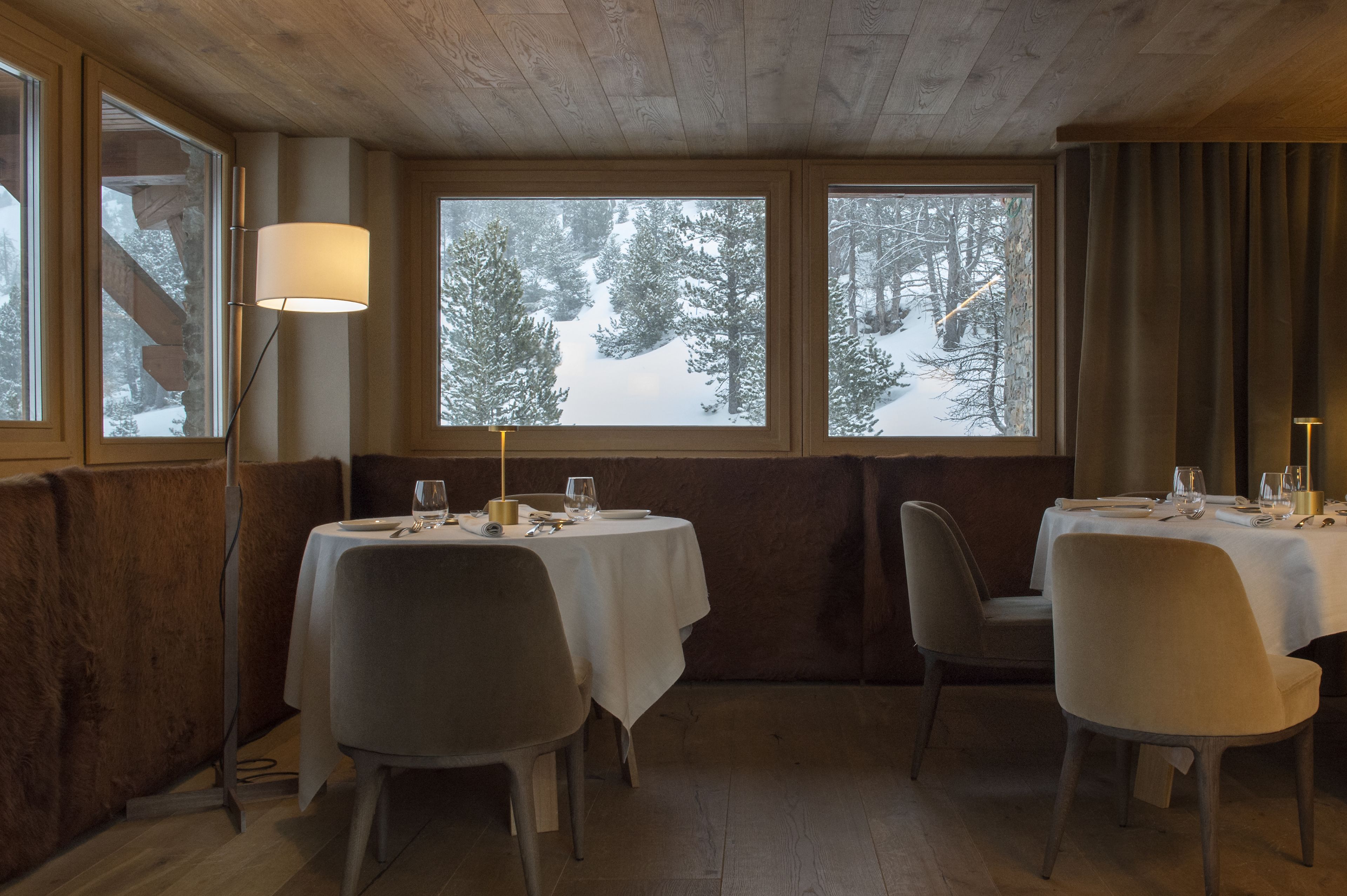 A cozy dining area with wooden walls and large windows overlooking a snowy forest. Tables are set with white tablecloths and chairs.