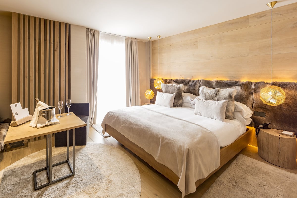 A modern hotel room with a large bed, a wooden headboard, a small table with a bottle and glasses, and a fur-covered bench.