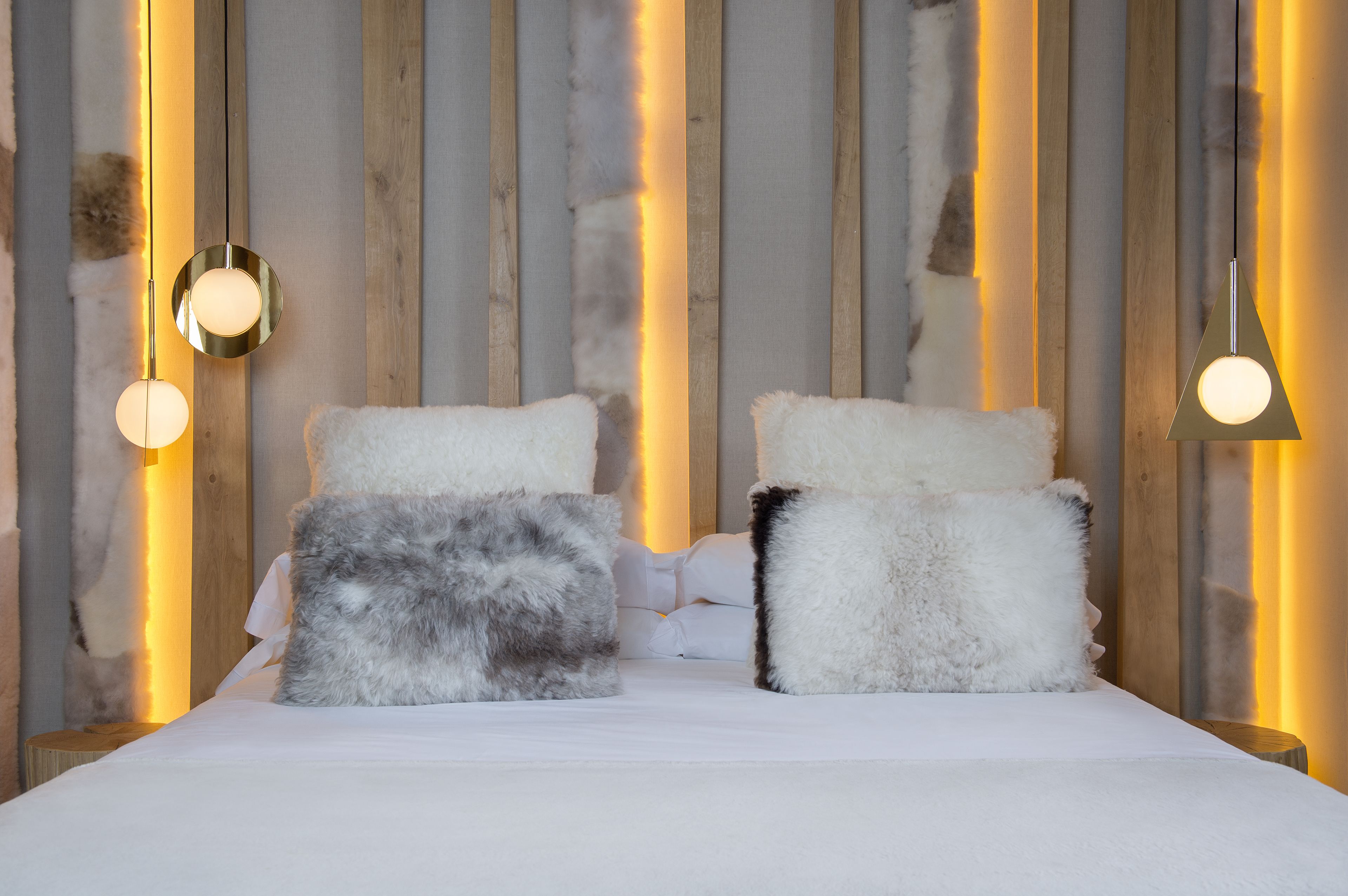 A bed with white bedding and fluffy pillows against a wooden wall with vertical light strips.