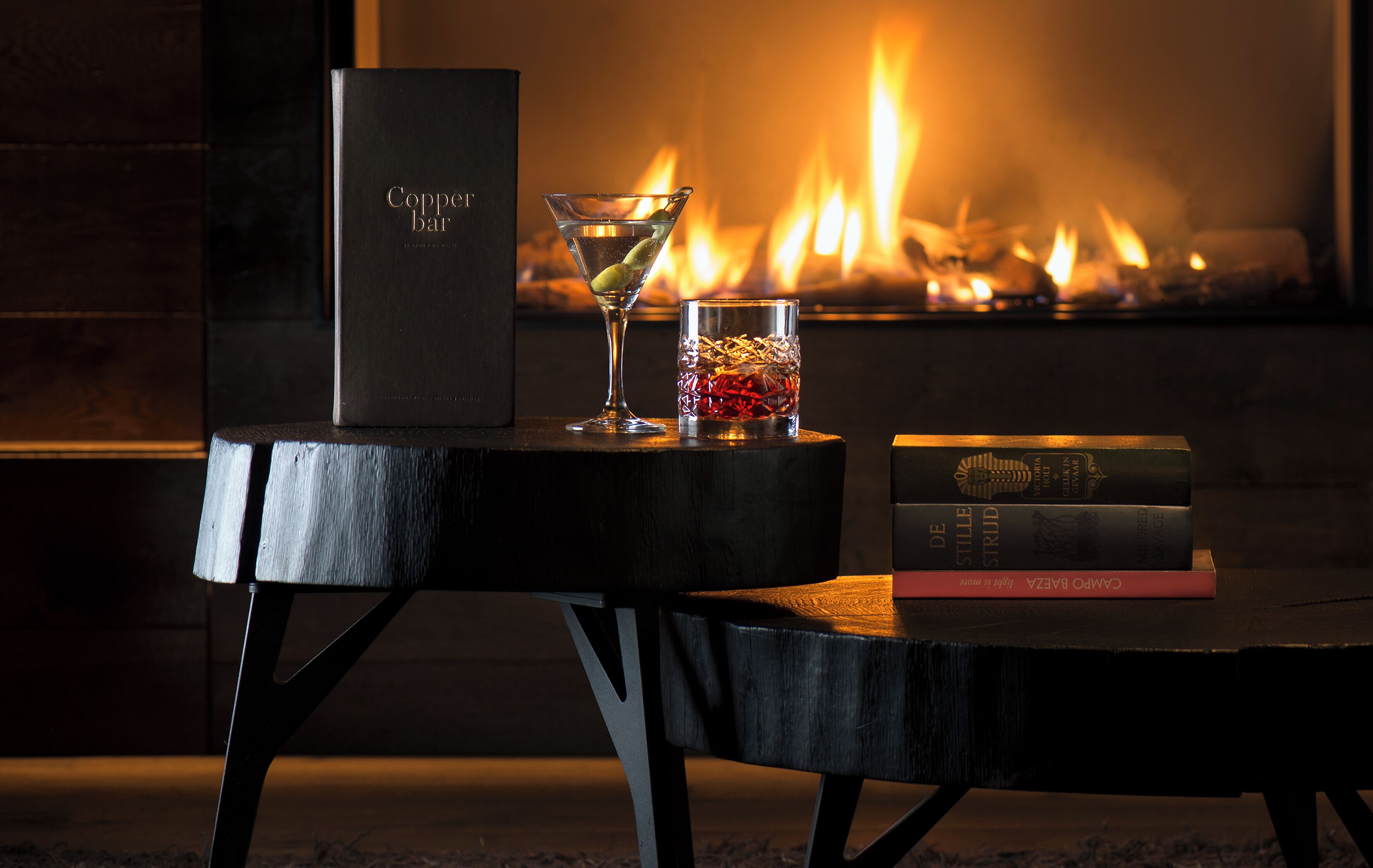 A table with a book titled "Carmen de la Paz" and a martini glass with a lime wedge, set against a fireplace with a lit fire.
