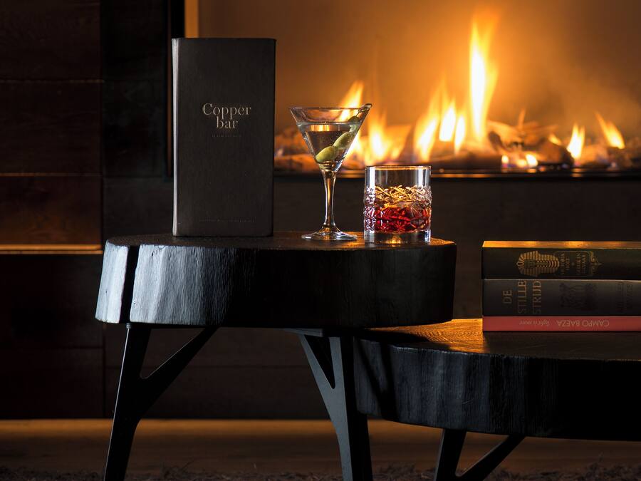 A table with a book titled "Carmen de la Paz" and a martini glass with a lime wedge, set against a fireplace with a lit fire.