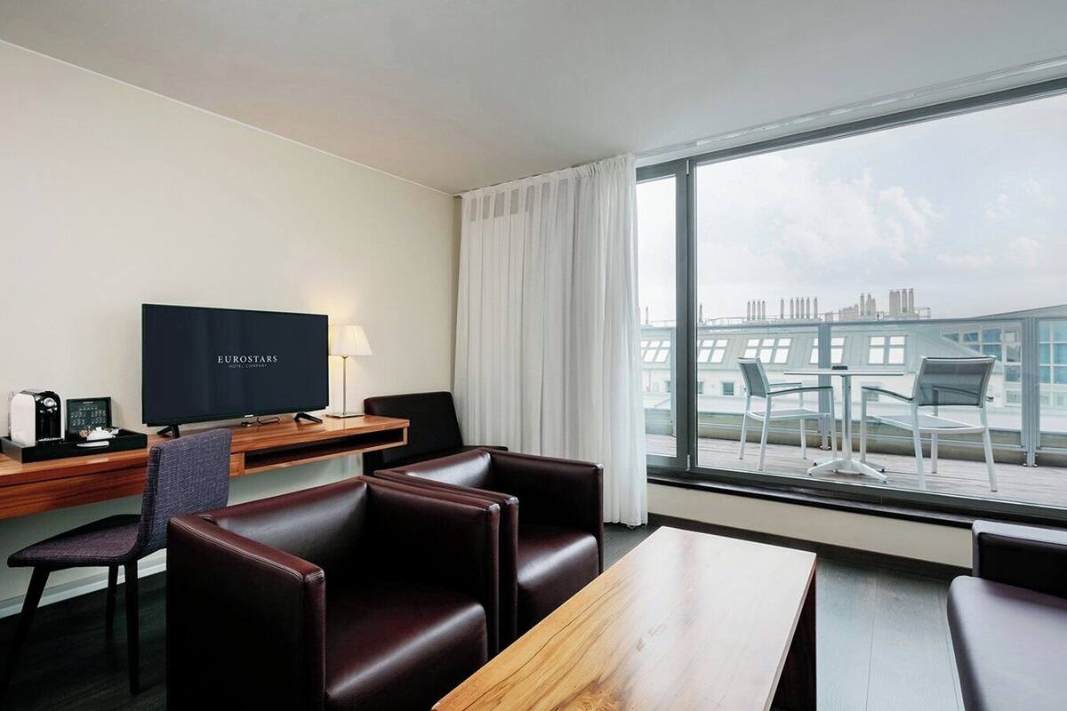 A hotel room with a large window, a flat-screen TV, leather seating, a wooden desk, and a view of the cityscape.