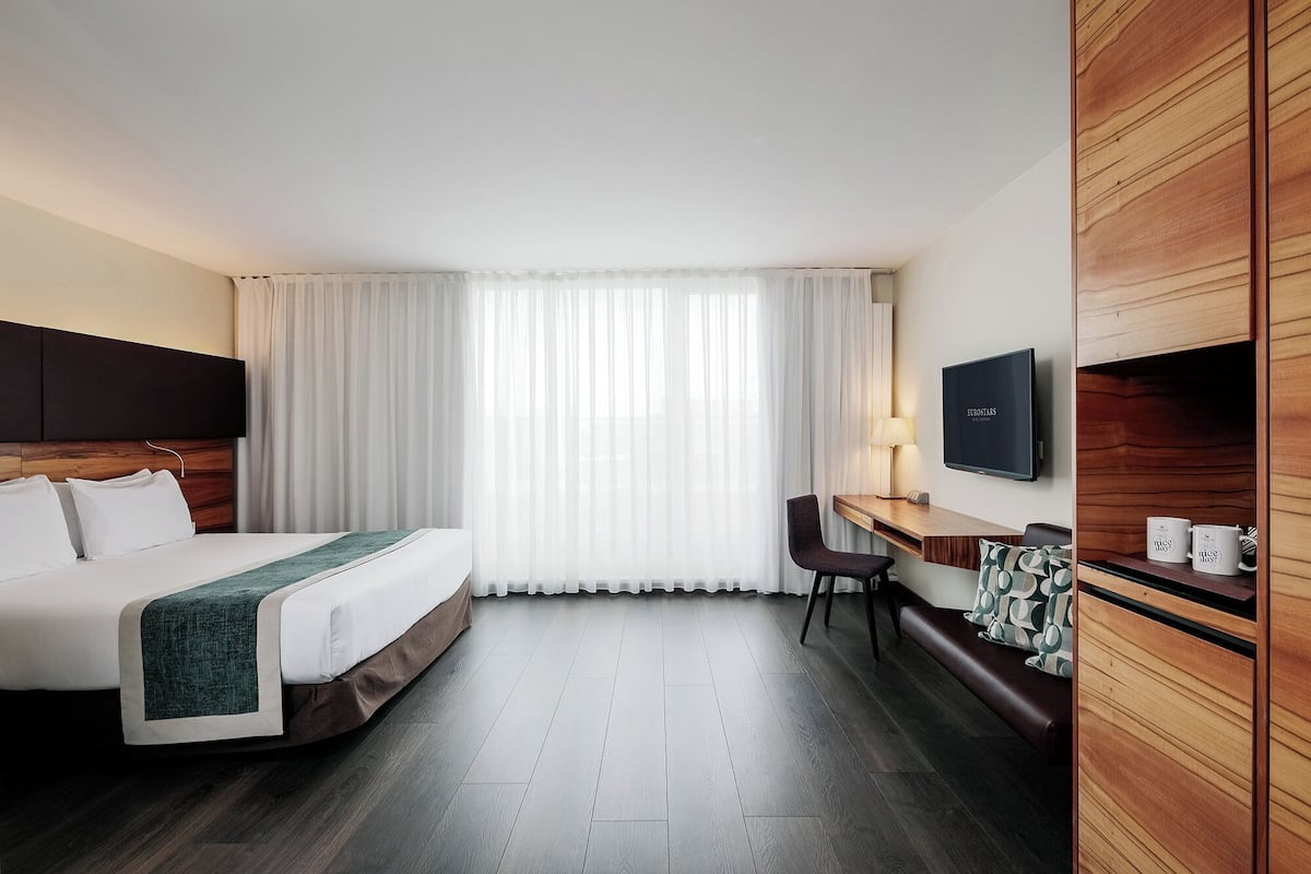 Una habitación de hotel con cama, escritorio, silla, televisión y ventana con cortinas.