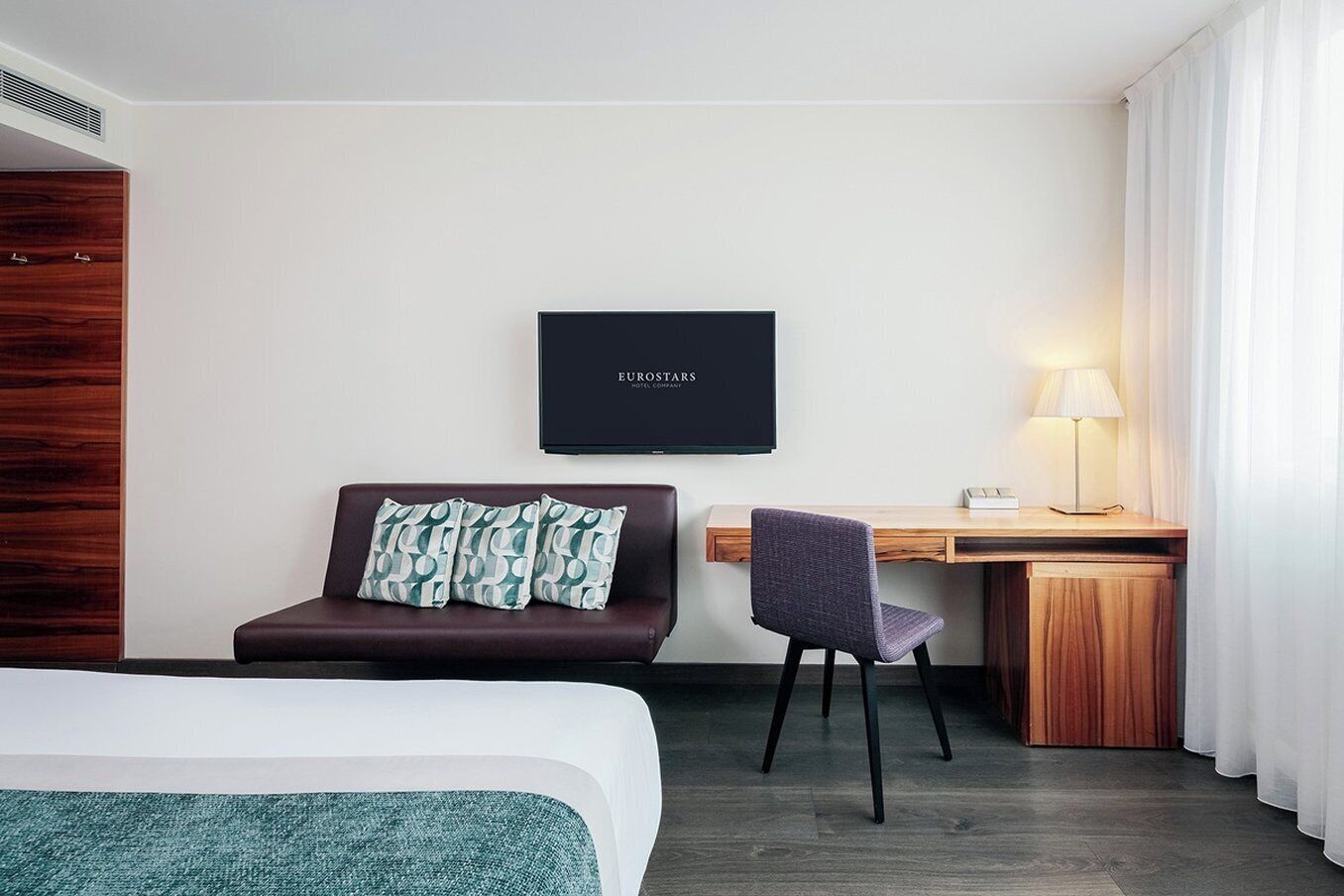 A hotel room with a bed, a desk with a chair, a television, and a window with curtains.
