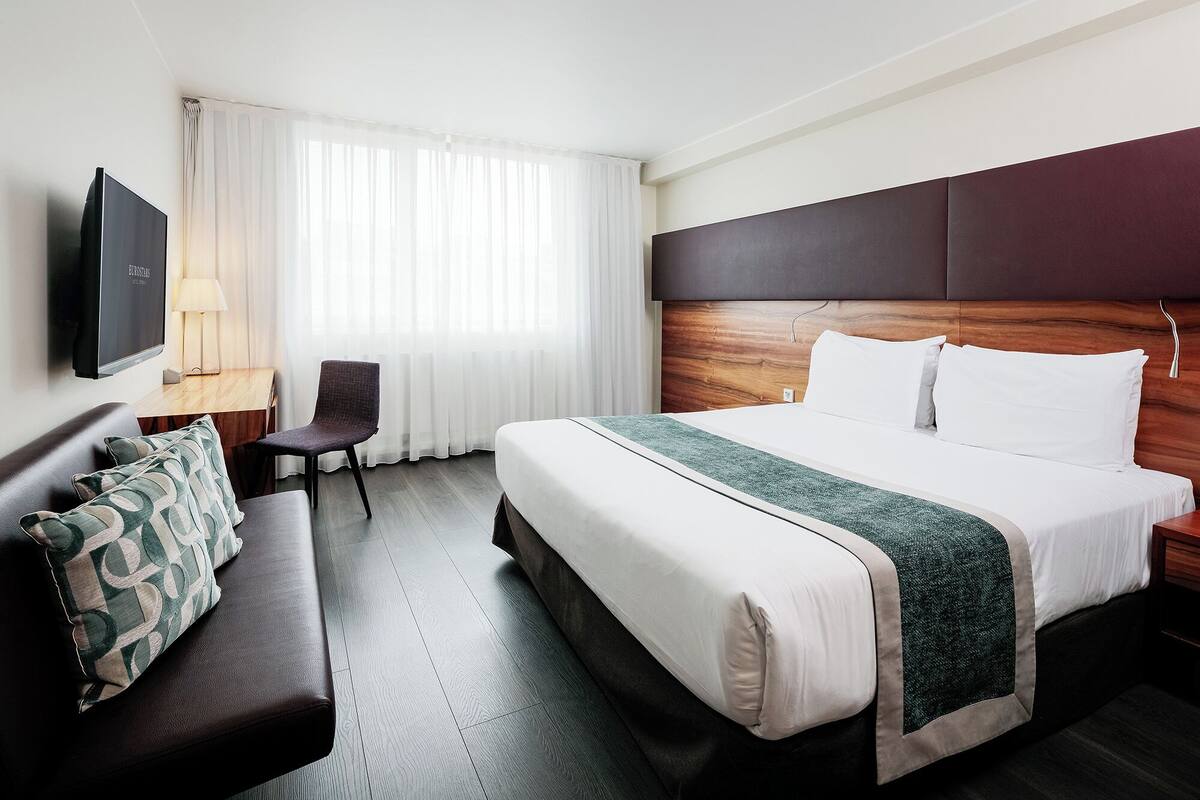 A hotel room with a large bed, a desk with a chair, a TV mounted on the wall, and a window with sheer curtains.