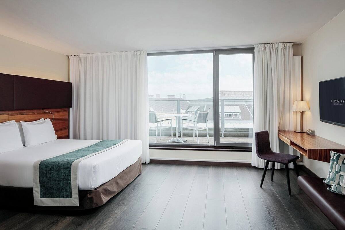 Habitación de hotel con cama, escritorio y silla, televisión y balcón con mesa y sillas.